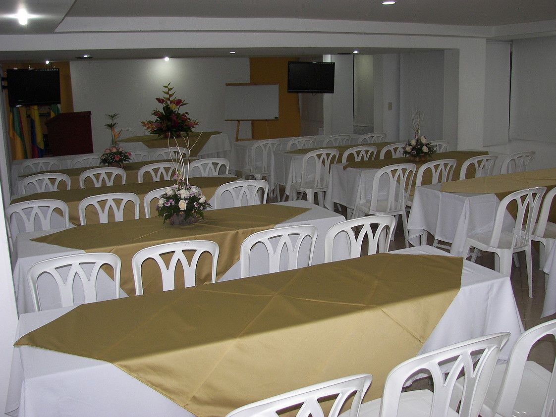 A banquet hall with tables covered in white tablecloths and gold runners, surrounded by white plastic chairs.