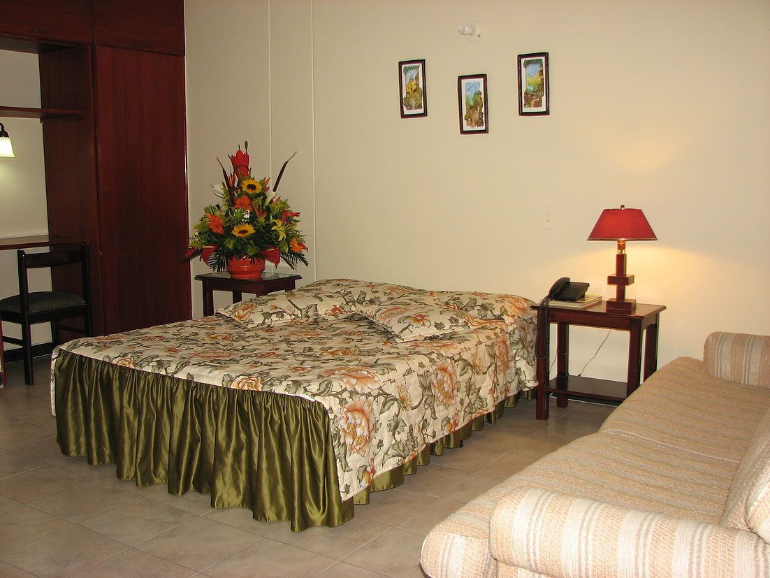 A bedroom with a bed, a bedside table with a lamp, a chair, a sofa, and a floral arrangement.