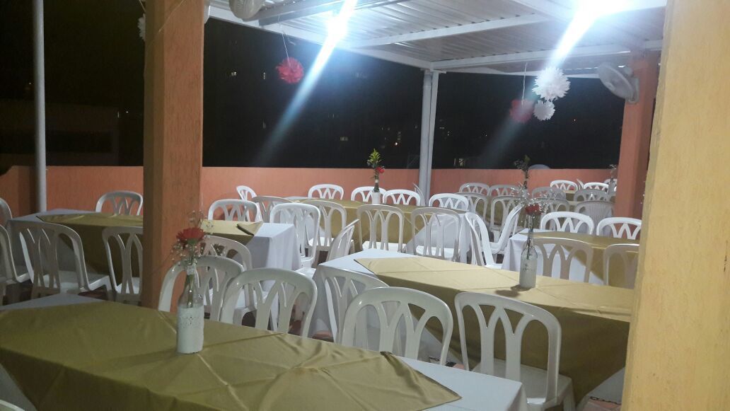 An outdoor dining area with tables and white plastic chairs, arranged under a covered section with a peach-colored wall and a dark night sky in the background.