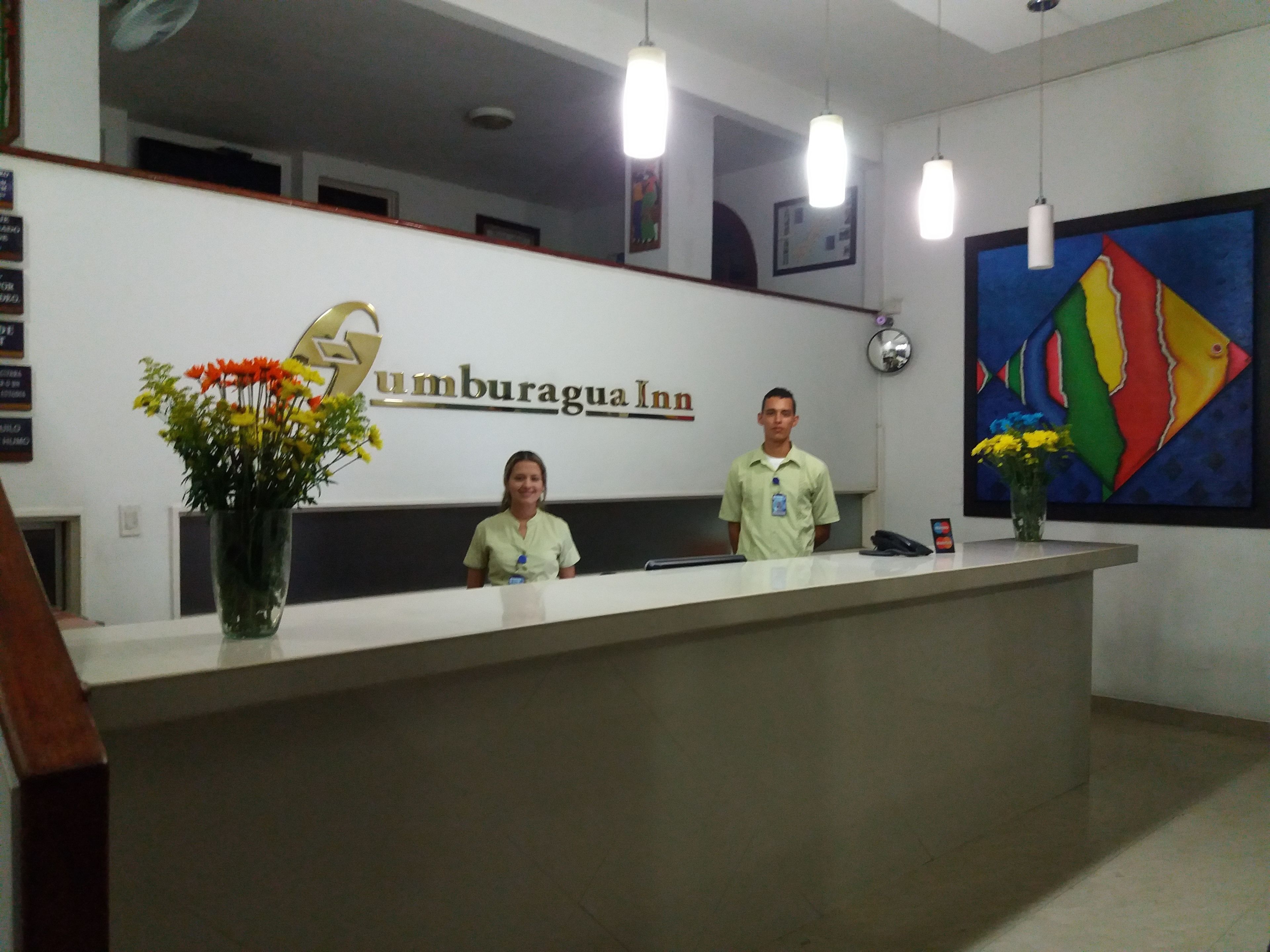A reception desk at Umburagua Inn with two staff members, a vase with flowers, and a colorful painting on the wall.