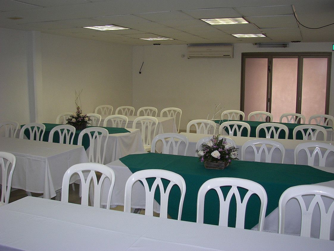A conference room with white plastic chairs and tables, each covered with a green tablecloth. There are floral centerpieces on the tables.