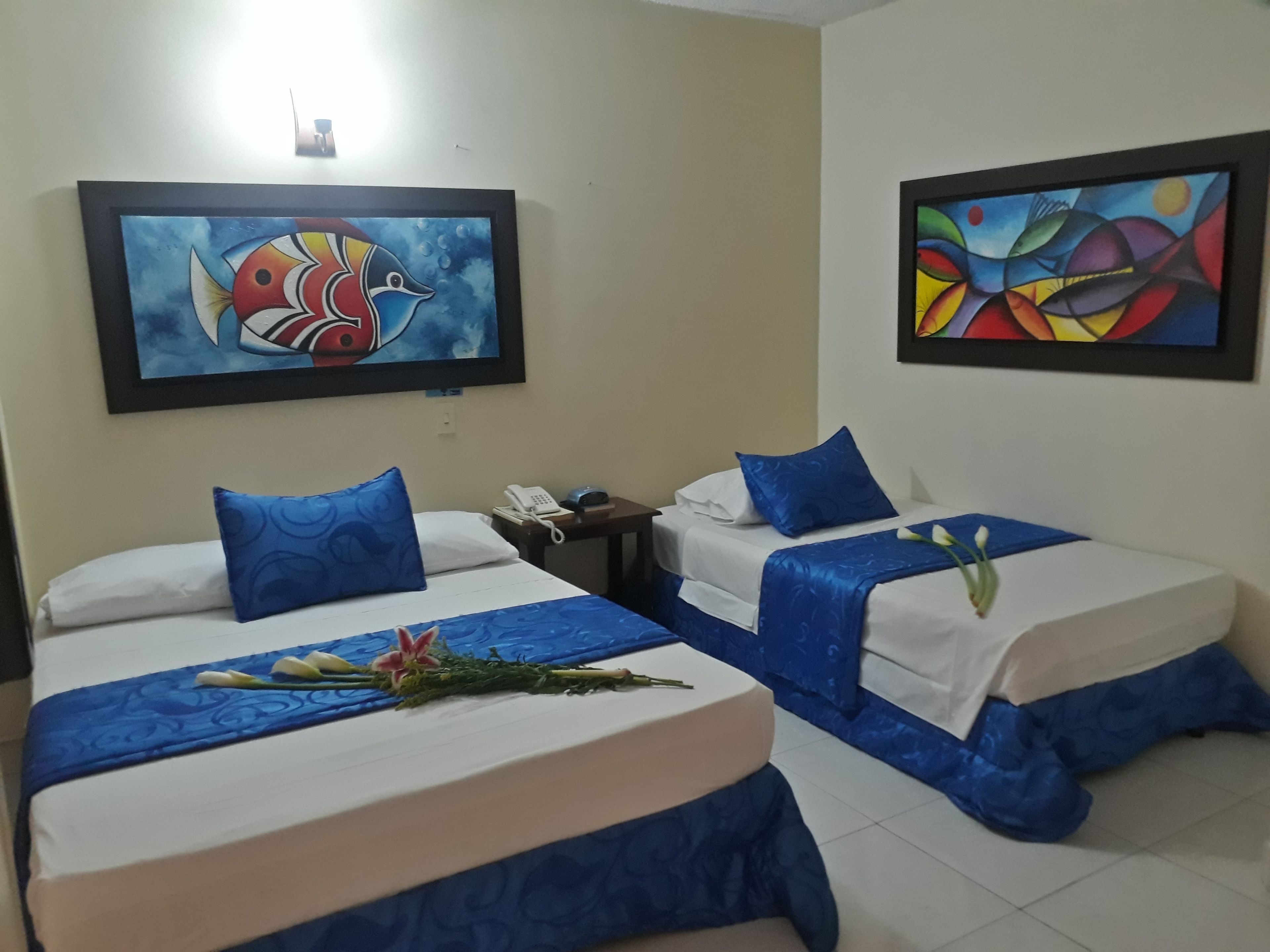 Habitación de hotel con dos camas, cada una con ropa de cama y almohadas azules, una mesita de noche con teléfono, y dos cuadros enmarcados en las paredes.