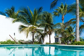 A swimming pool with clear blue water, surrounded by palm trees and a white building. - Cas Elizabeth (Noord)