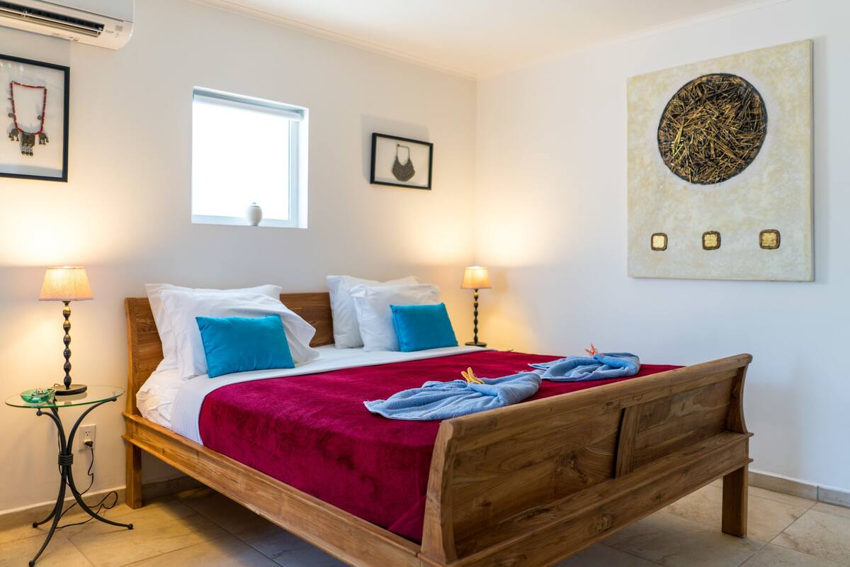 En snyggt bäddad säng med ett rött överkast, två kuddar och en sänggavel i trä. Det finns två sängbord med lampor, ett fönster med utsikt och ett konstverk som hänger på väggen.