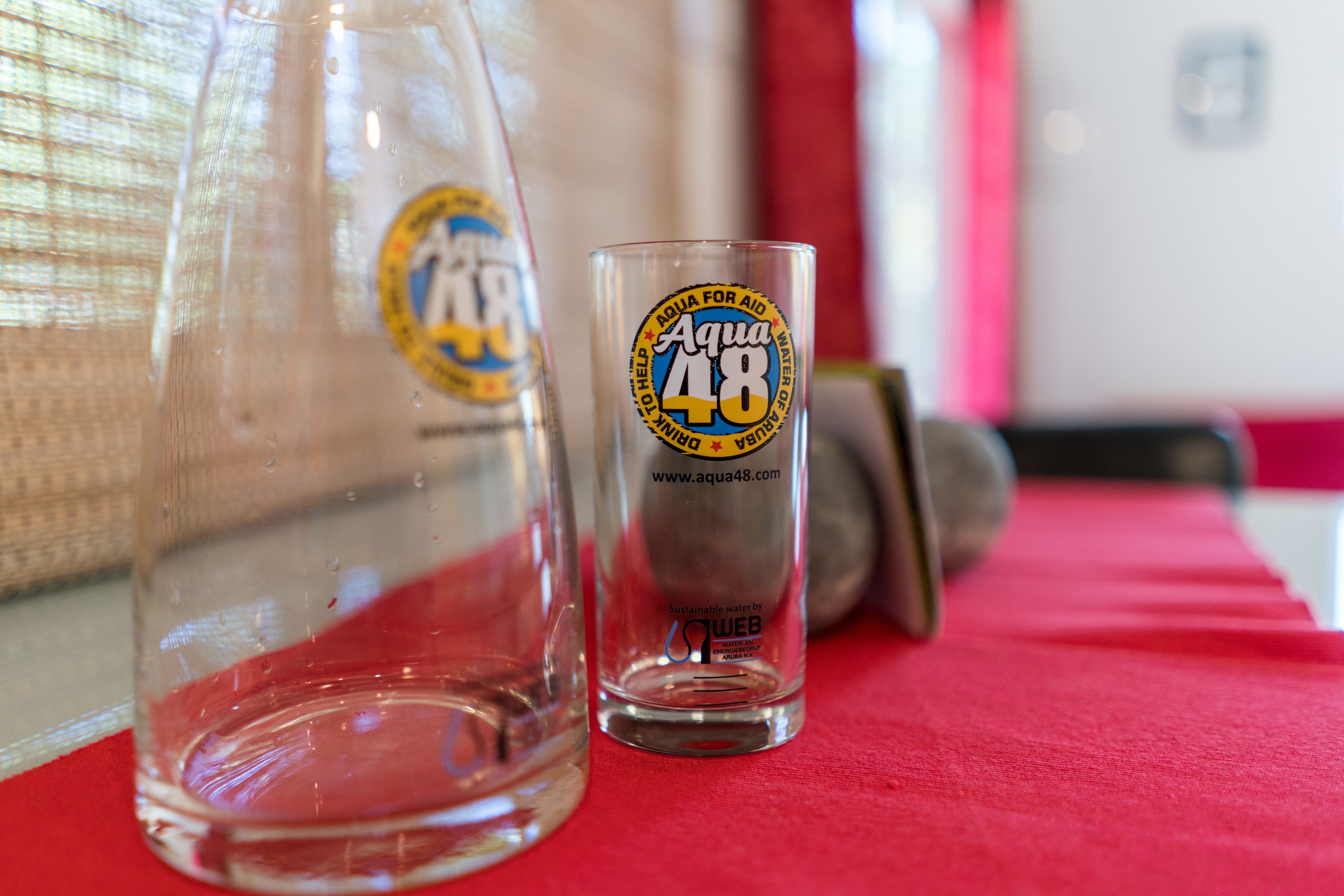 A clear glass bottle and a glass with the Aqua 48 logo on a red surface.