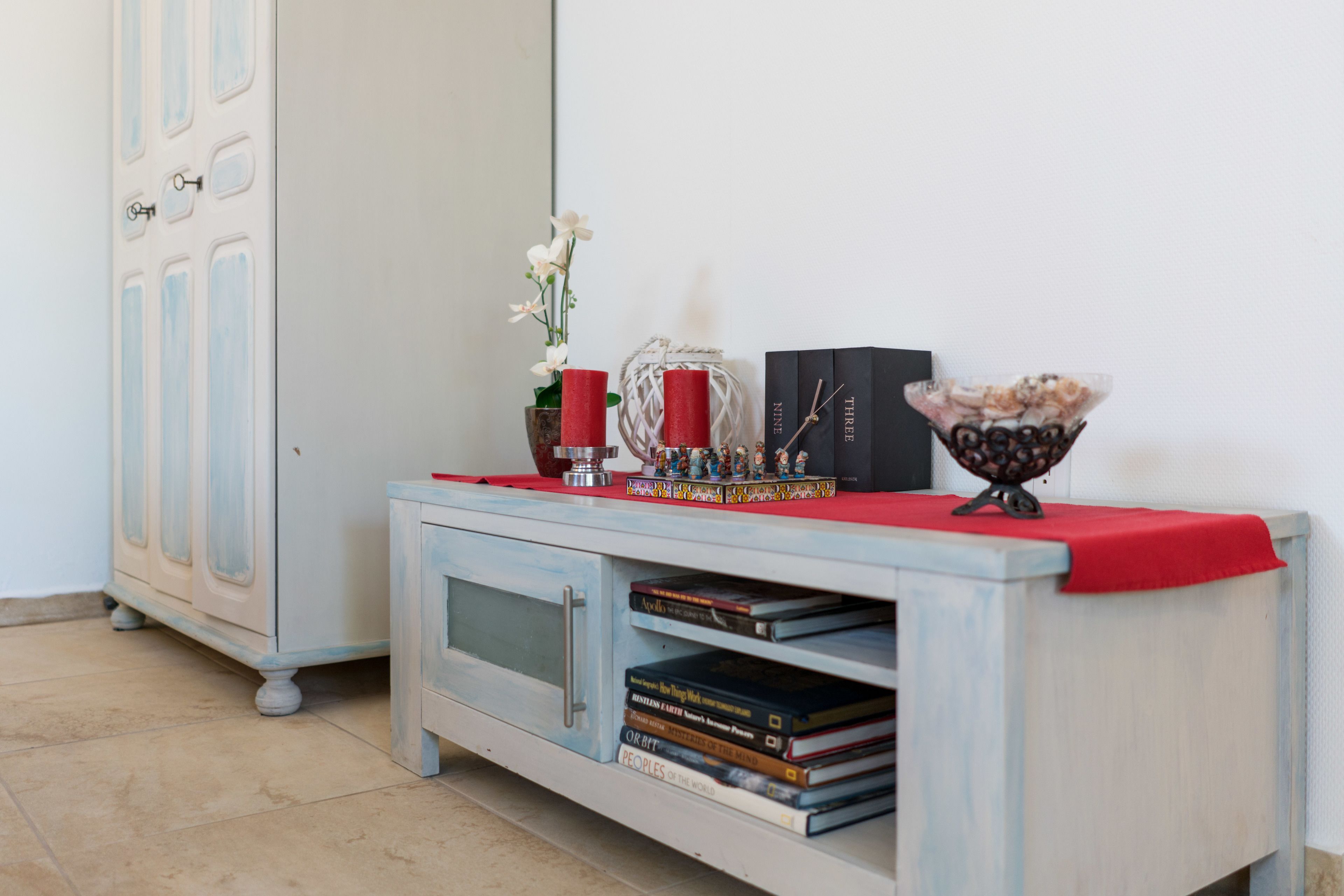 A white cabinet with a red cloth, books, a red candle, a white flower, and a decorative item on top.