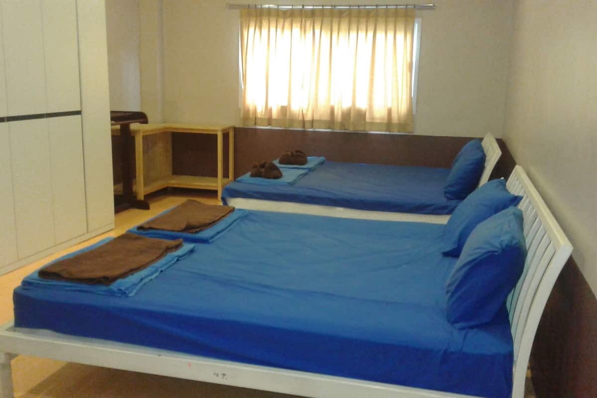 A room with two beds, each with blue bedding and pillows, a small bedside table with a lamp, and a window with curtains.