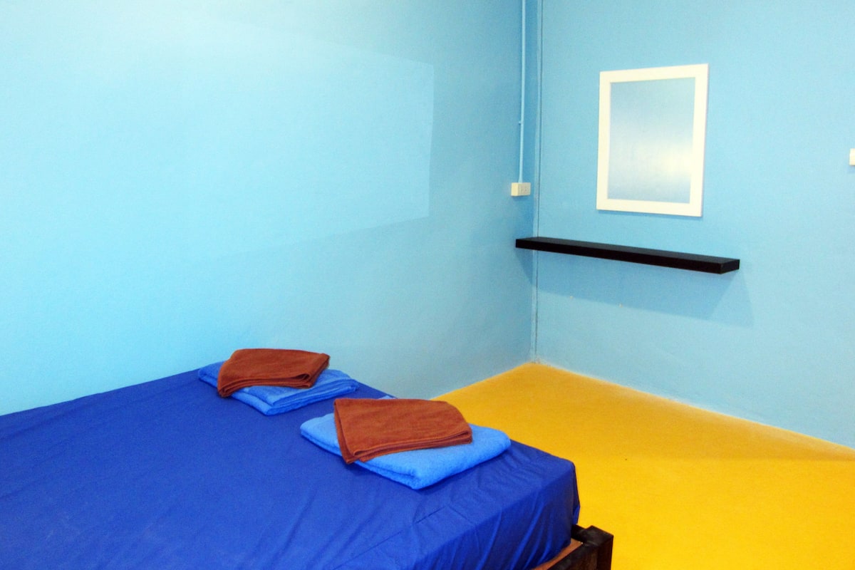 A room with two beds, one with blue bedding and the other with yellow bedding. There is a black shelf on the wall and a white framed picture above it.