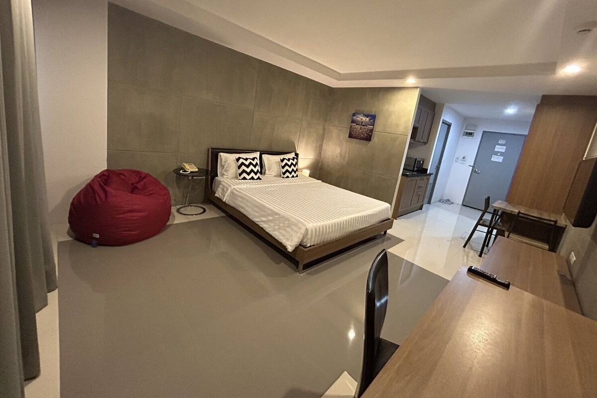 Una habitación de hotel moderna con una cama grande, un sillón de peluche rojo, una mesita de noche con un teléfono y una kitchenette al fondo.
