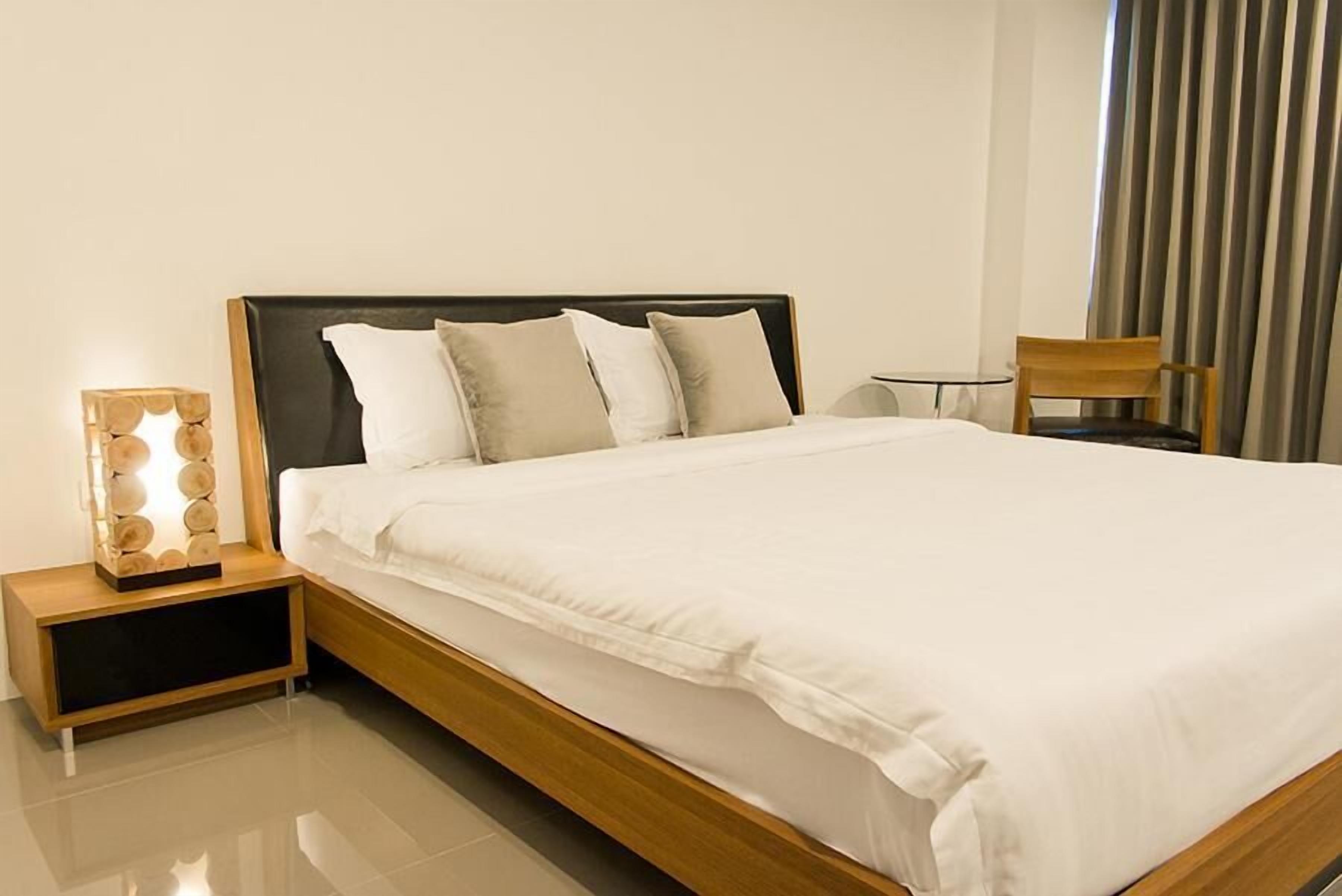 A neatly made bed with a wooden headboard and white bedding, a bedside table with a lamp, and a chair in a modern hotel room.