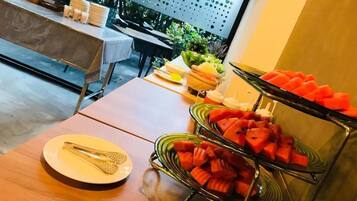 A buffet table with watermelon and other fruits, a table with plates and cutlery, and a view of the outdoors.