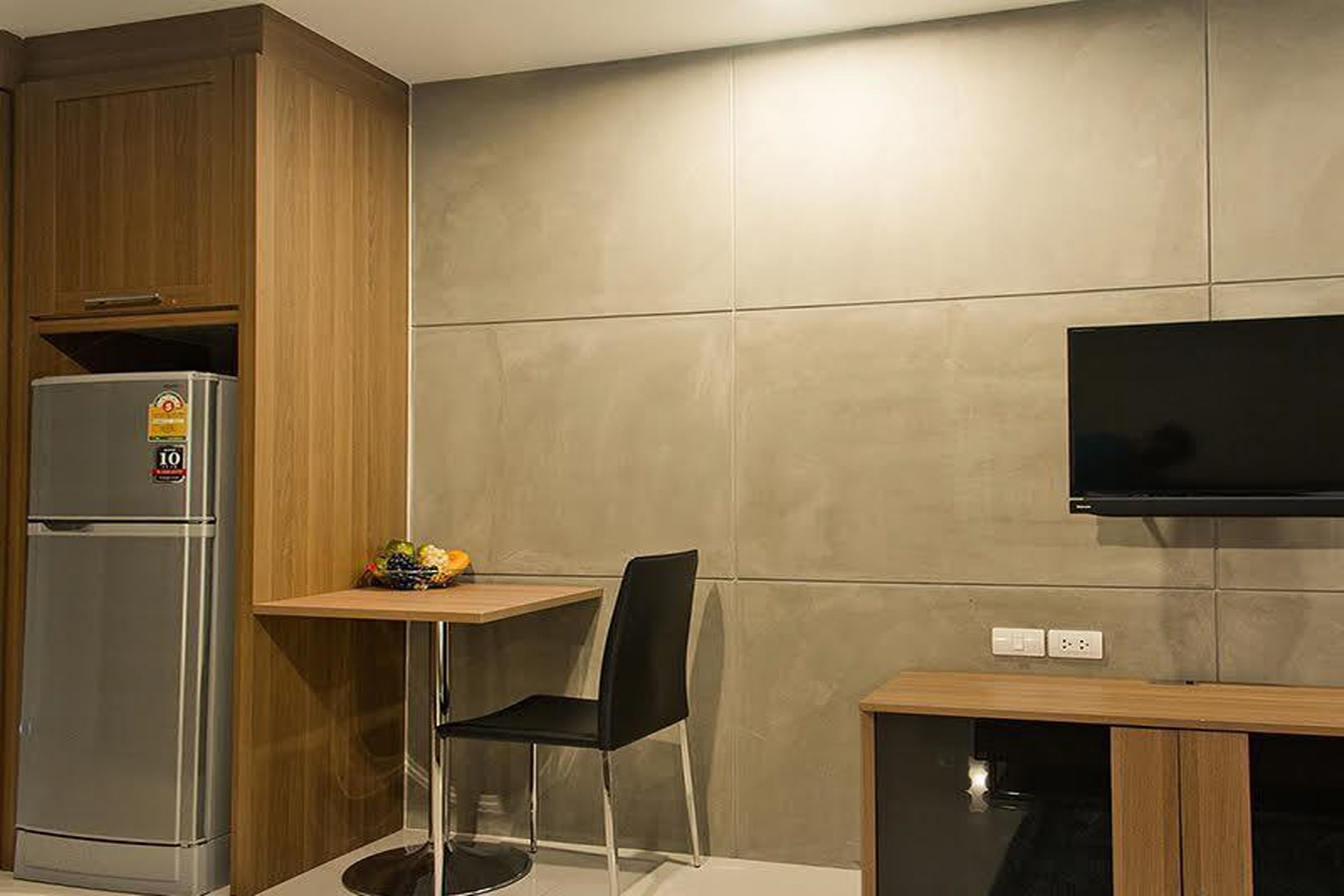 A compact kitchenette with a refrigerator, a small table with a chair, and a flat-screen TV mounted on the wall.