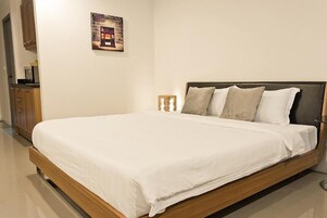 Una cama bien tendida con estructura de madera y ropa de cama blanca en una habitación con suelo de baldosas y un mueble de madera.