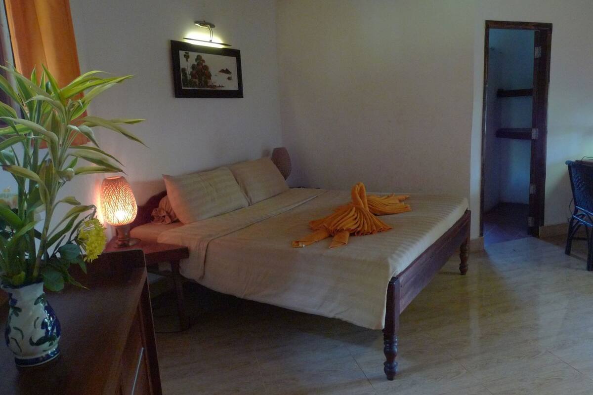 A room with a bed, a desk, a chair, a potted plant, and a wall-mounted picture.