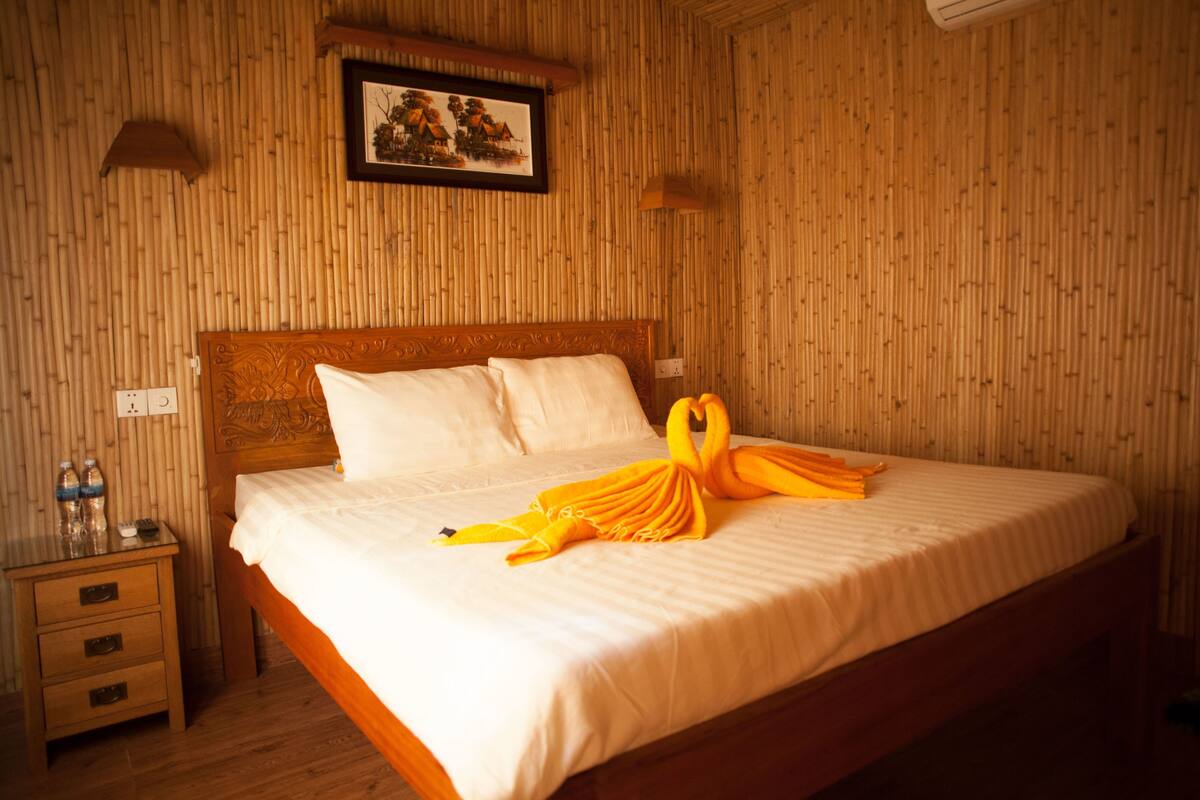 A neatly made bed with a swan towel arrangement, a bedside table with bottles, and a framed picture on the wall.