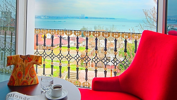 Une pièce avec une grande fenêtre, un fauteuil rouge, une table avec un journal, une tasse et un vase.