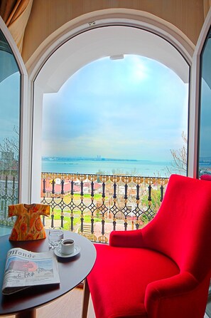 Une pièce avec une grande fenêtre, un fauteuil rouge, une table avec un journal, une tasse et un vase.