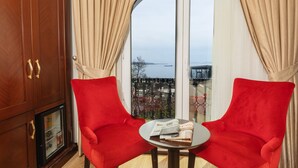 Une pièce avec deux chaises rouges, une petite table sur laquelle se trouve un magazine, un meuble en bois et une grande fenêtre donnant sur un plan d’eau.