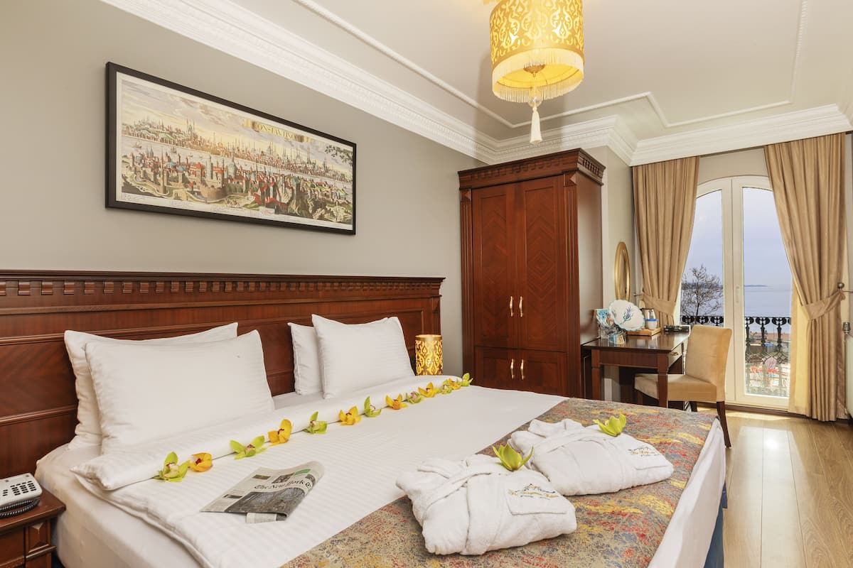 Ein Hotelzimmer mit einem großen Bett, einem hölzernen Kopfteil, einem gerahmten historischen Gemälde, einem Schreibtisch mit Lampe und Blick auf das Meer durch das Fenster.
