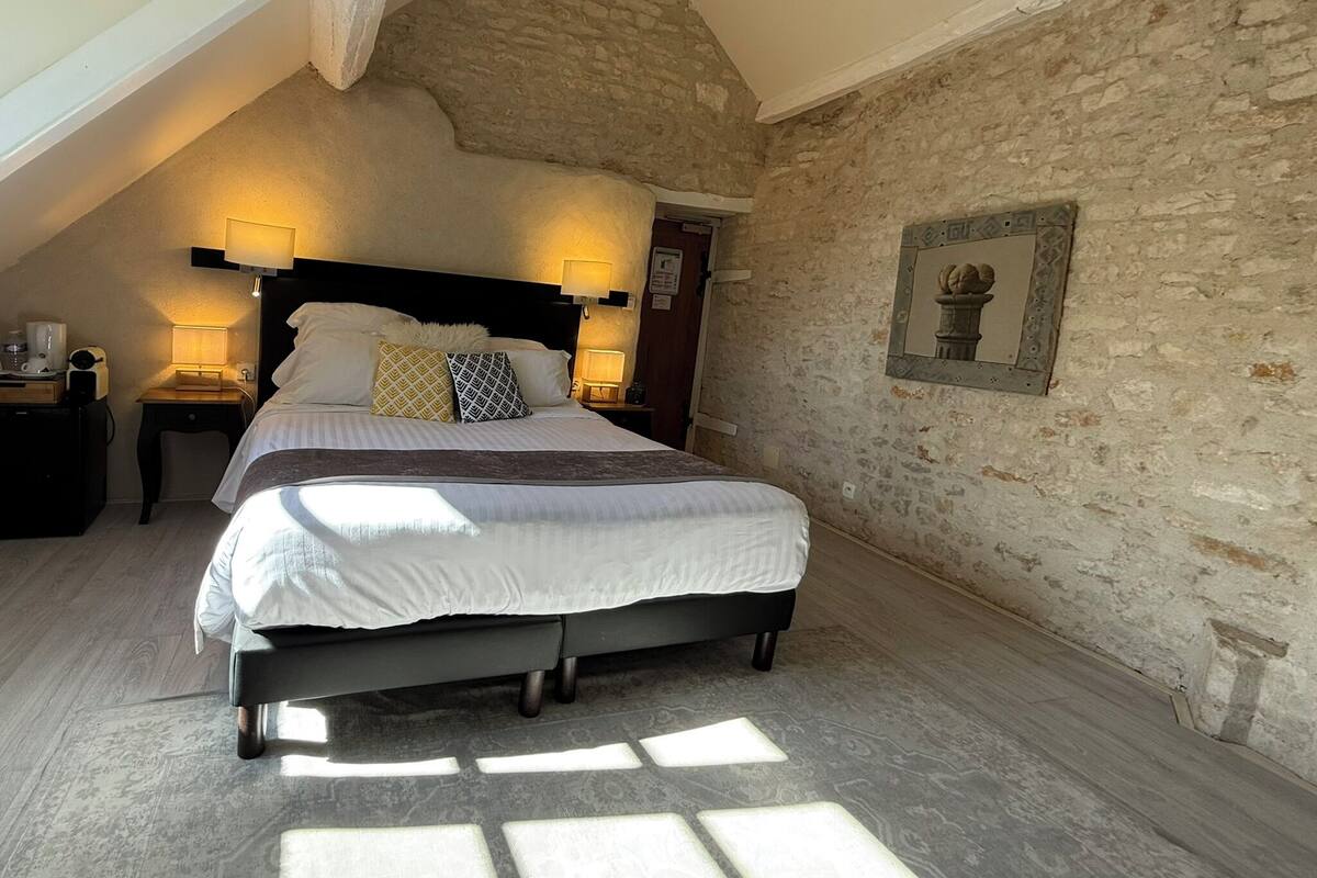 Une chambre avec un plafond en pente, un mur en pierre, un lit avec une literie blanche, deux lampes de chevet et un tableau encadré au mur.