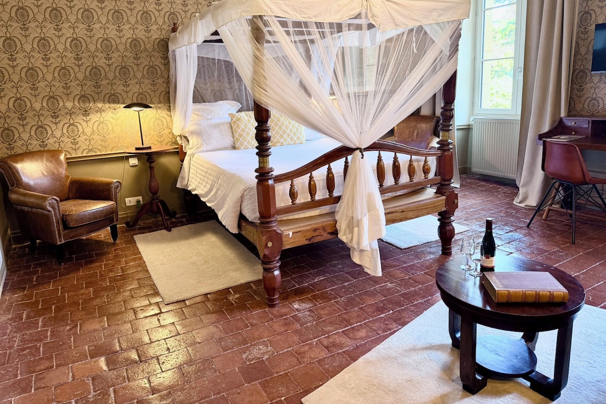 Un lit traditionnel à baldaquin, un fauteuil en cuir, une table de chevet avec une bouteille et des verres, et un bureau avec un livre.