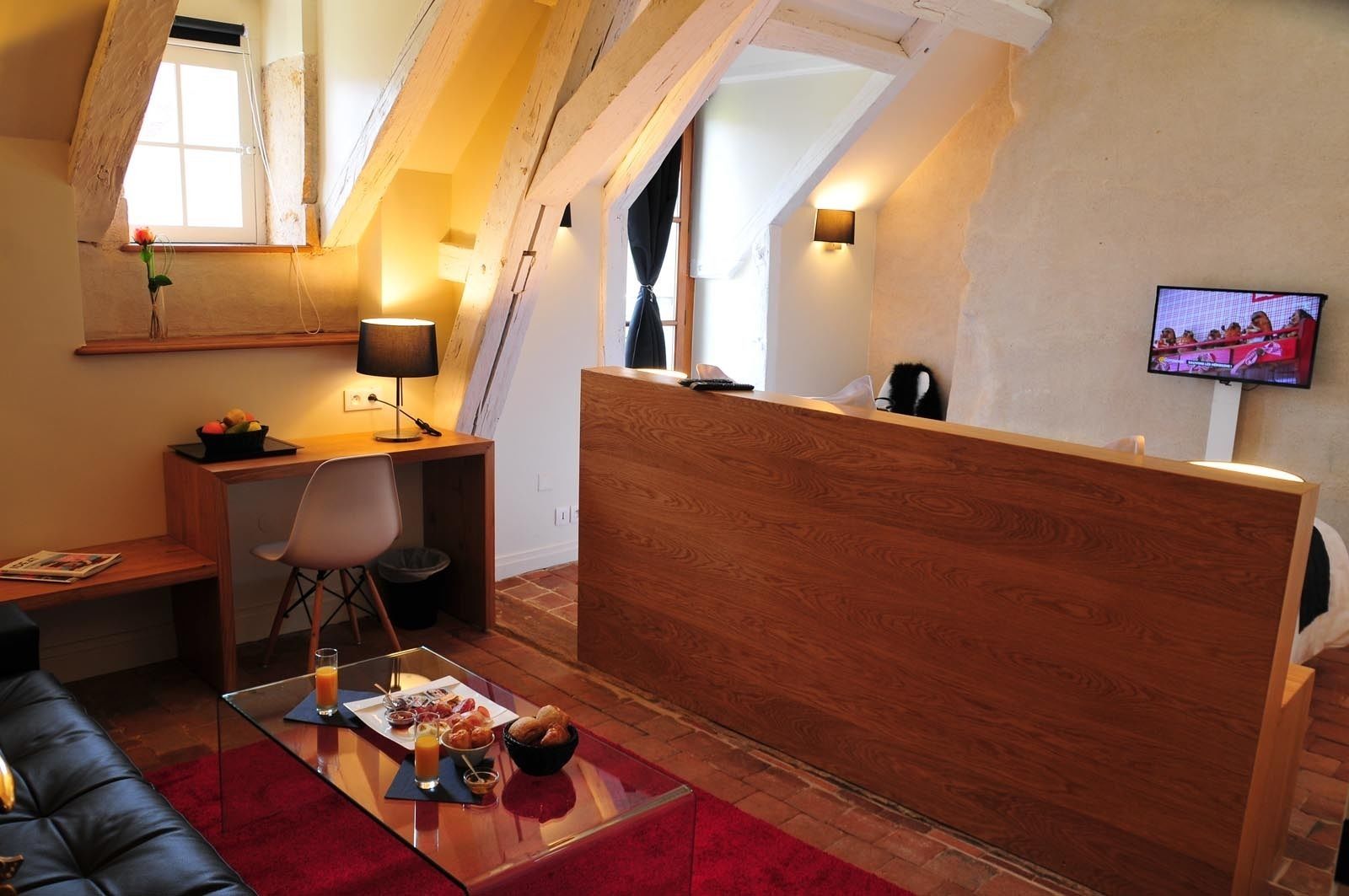Ein gemütliches Zimmer mit Schreibtisch aus Holz, einem Stuhl, einem Glastisch mit Frühstücksangeboten, einem roten Teppich und einem Fernseher an der Wand.