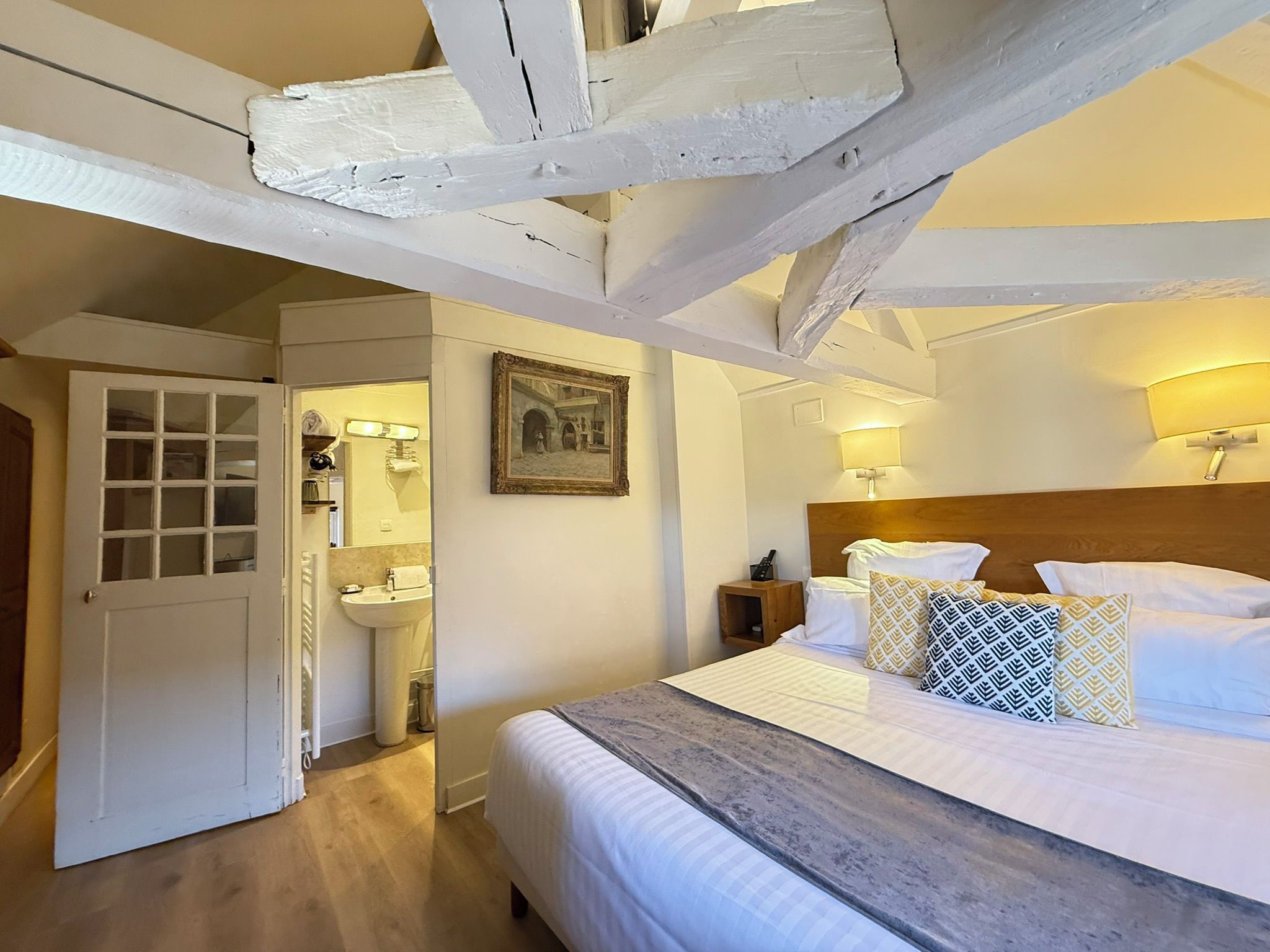 A bedroom with a bed, bedside lamps, a wooden headboard, a painting, and a door leading to a bathroom.