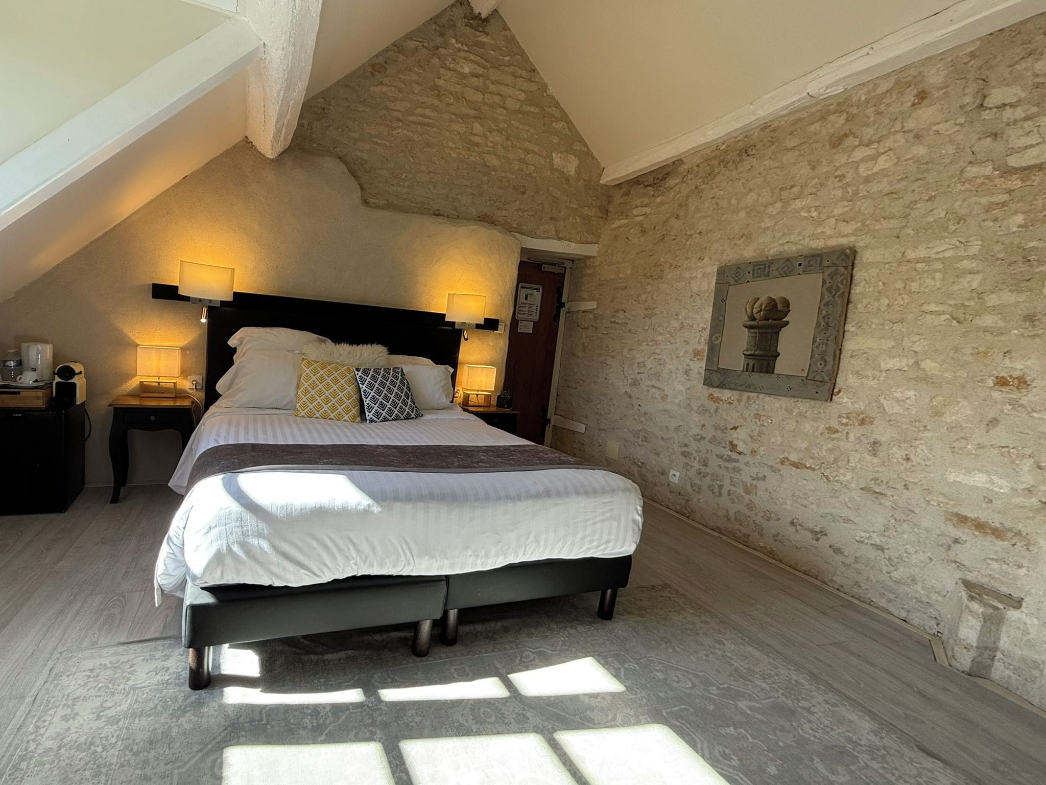 A bedroom with a sloped ceiling, a stone wall, a bed with white bedding, two bedside lamps, and a framed picture on the wall.