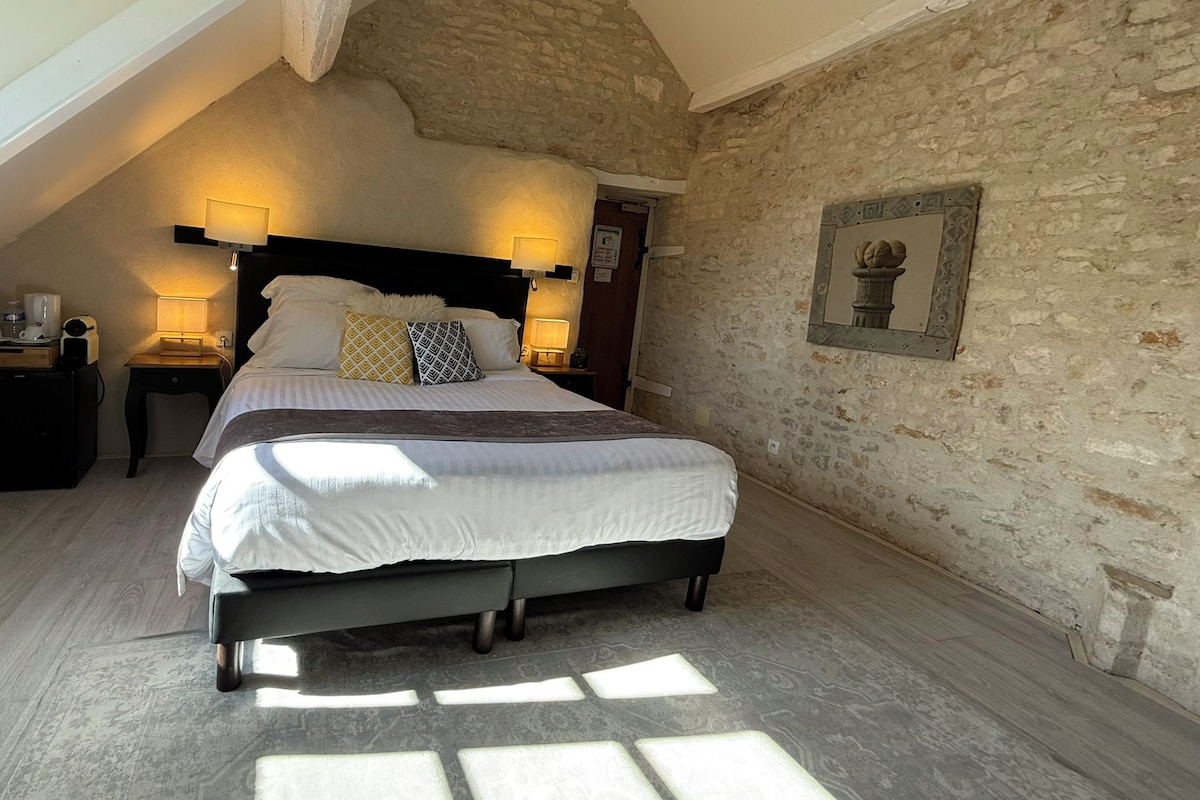 Une chambre avec un plafond en pente, un mur en pierre, un lit avec une literie blanche, deux lampes de chevet et un tableau encadré au mur.