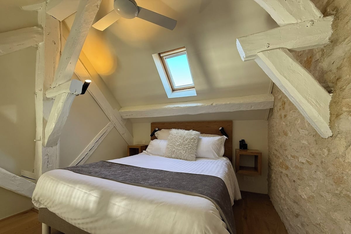 Une chambre mansardée chaleureuse, avec un plafond en pente, un puits de lumière, une tête de lit en bois et un lit recouvert de draps blancs et agrémenté d’un oreiller moelleux.