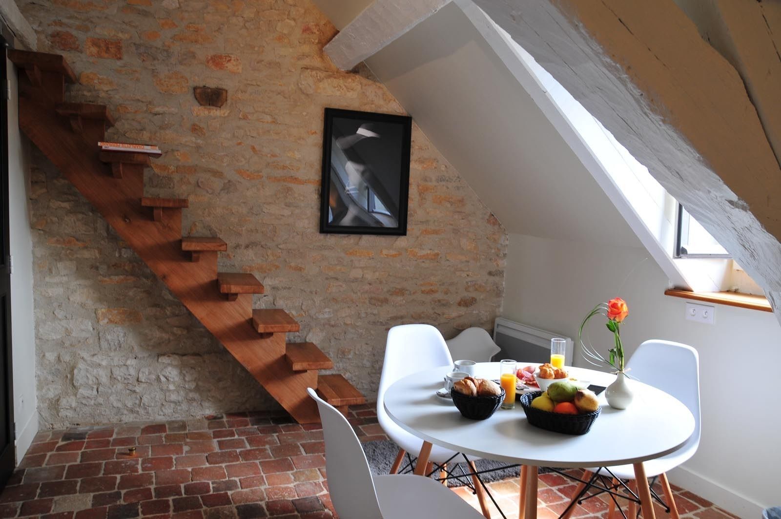 A dining area with a brick floor, white table, and chairs. A staircase with wooden steps leads to a lofted area.