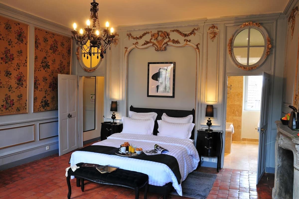 Une chambre spacieuse avec un grand lit, un lustre, une cheminée et une vue sur l’extérieur.