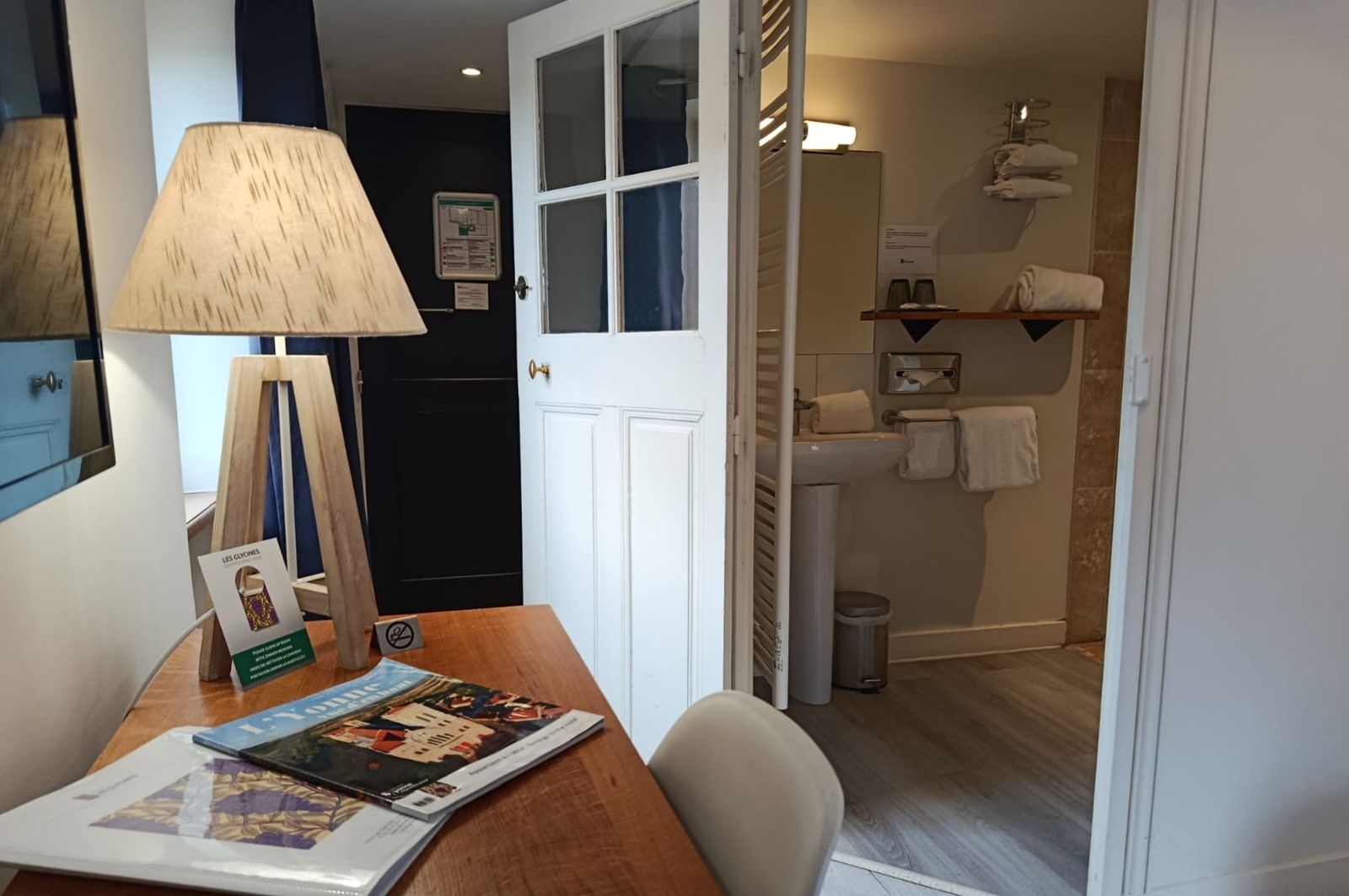 A room with a wooden desk, a lamp, and a door leading to a bathroom with a sink and towel rack.