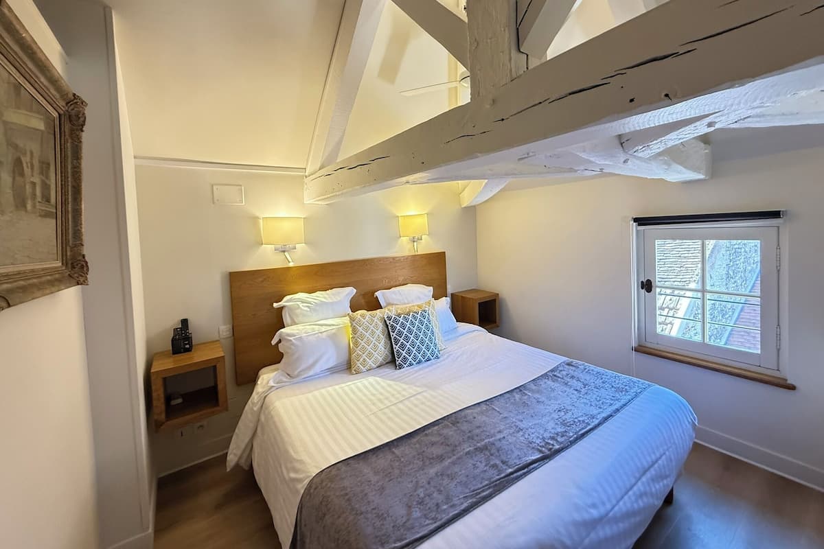 Une chambre dotée d’un grand lit, de deux tables de chevet, de deux lampes fixées au mur, d’une fenêtre offrant une vue agréable et de poutres apparentes.