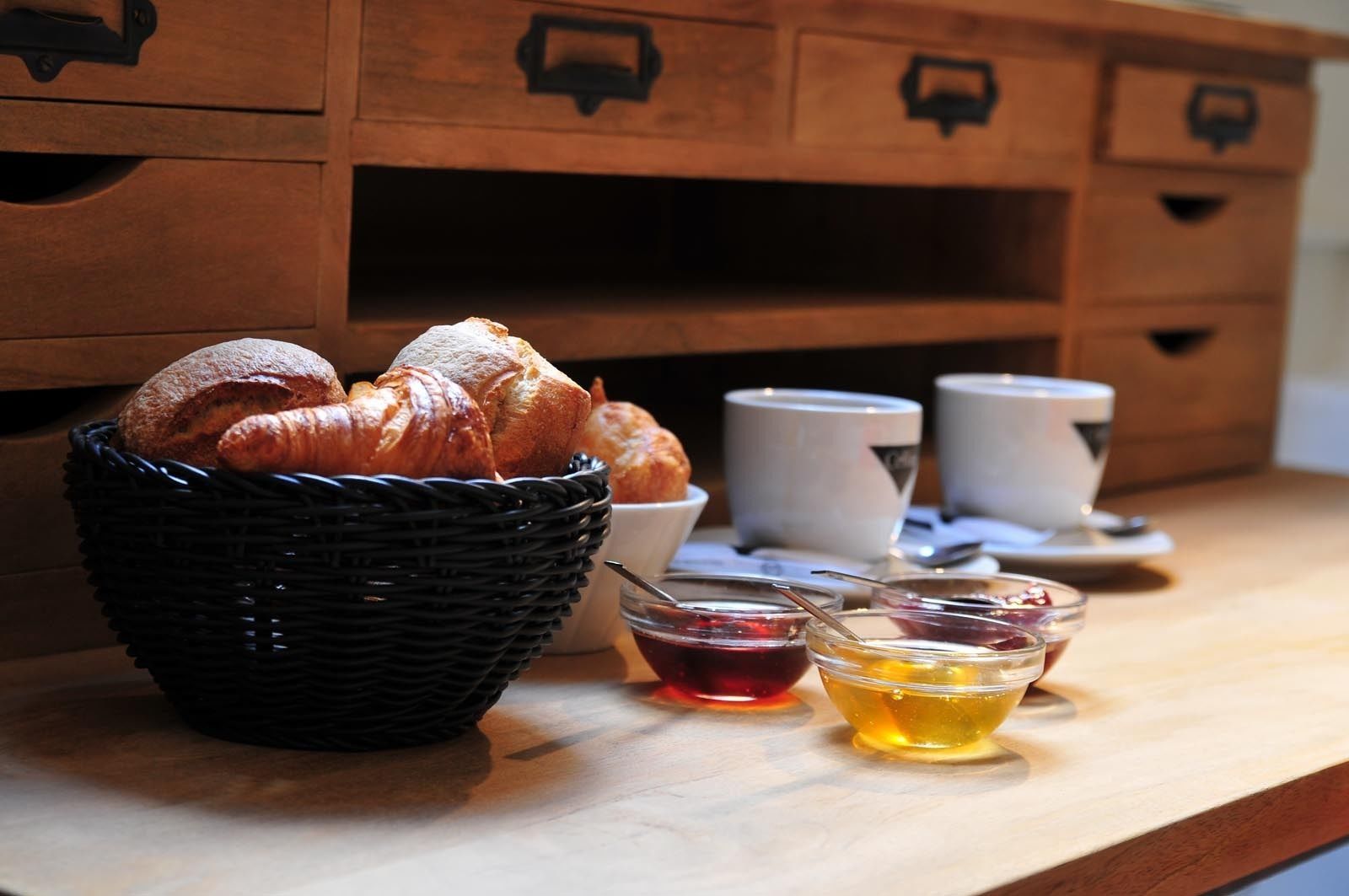 Uma cesta de croissants sobre uma mesa de madeira, com duas pequenas tigelas de compotas e mel.