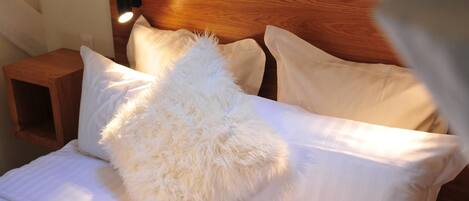 A neatly made bed with white bedding and fluffy pillows, a wooden headboard, and a bedside table with a lamp.
