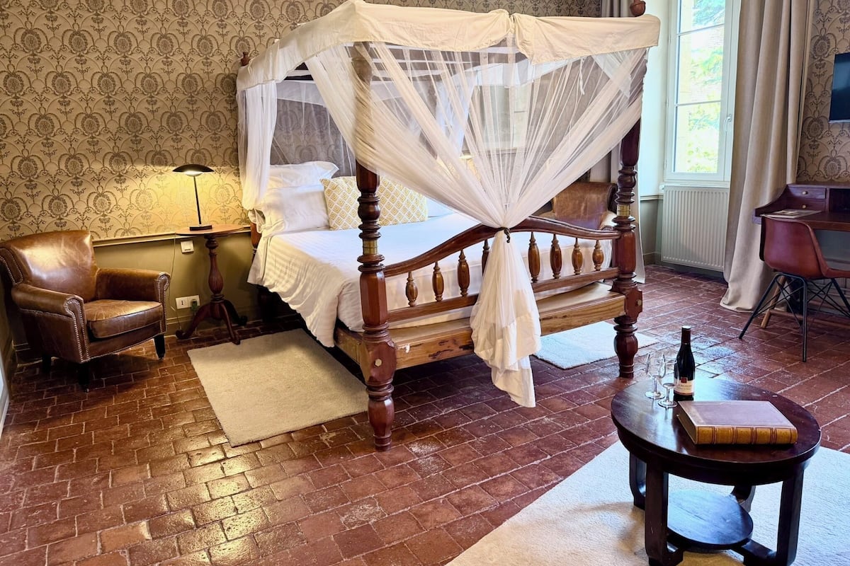 Un lit traditionnel à baldaquin, un fauteuil en cuir, une table de chevet avec une bouteille et des verres, et un bureau avec un livre.