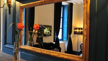 Un espejo con marco dorado que refleja una habitación con puerta azul, un cuadro y una lámpara.