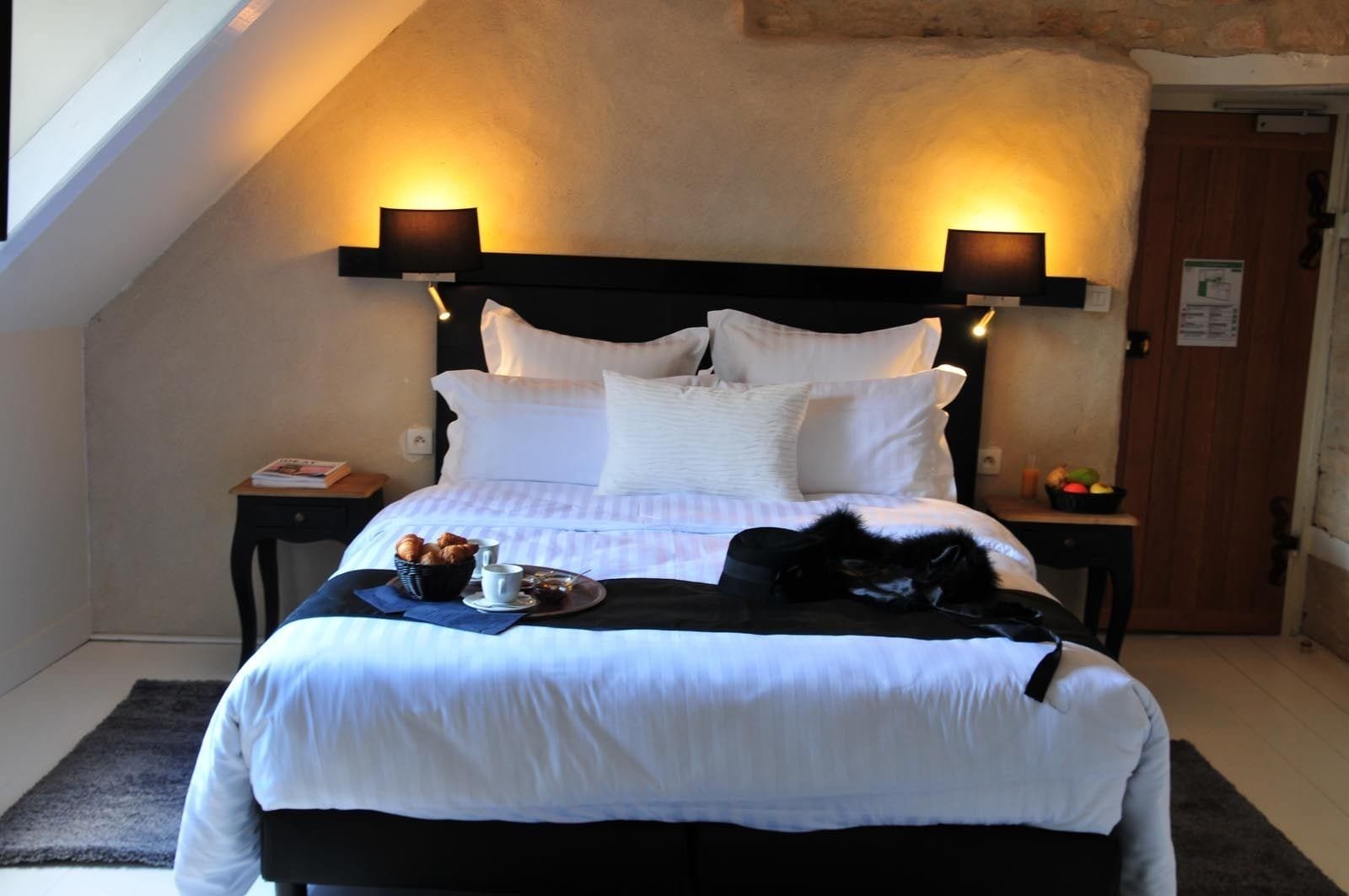 A neatly made bed with white linens, a black headboard, and two bedside lamps. There is a tray with croissants and a cup on the bed.
