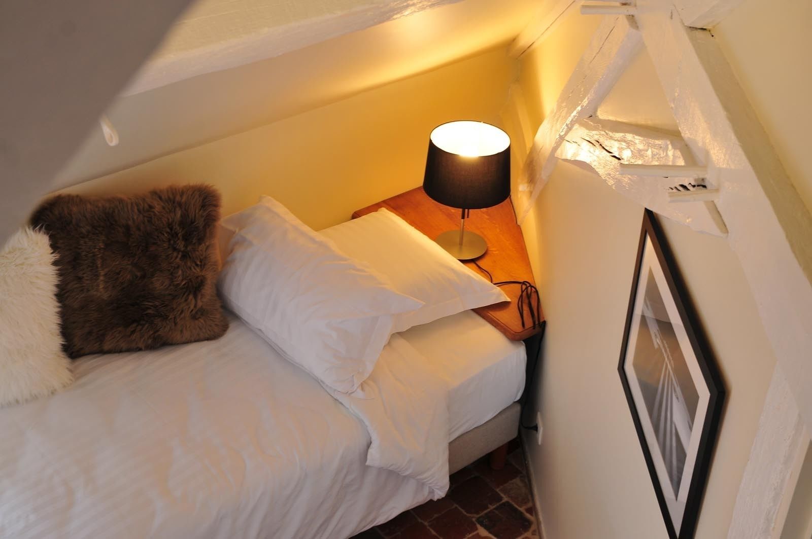 Et hyggeligt, lille værelse med en seng, en lampe og et billede i ramme på væggen.
