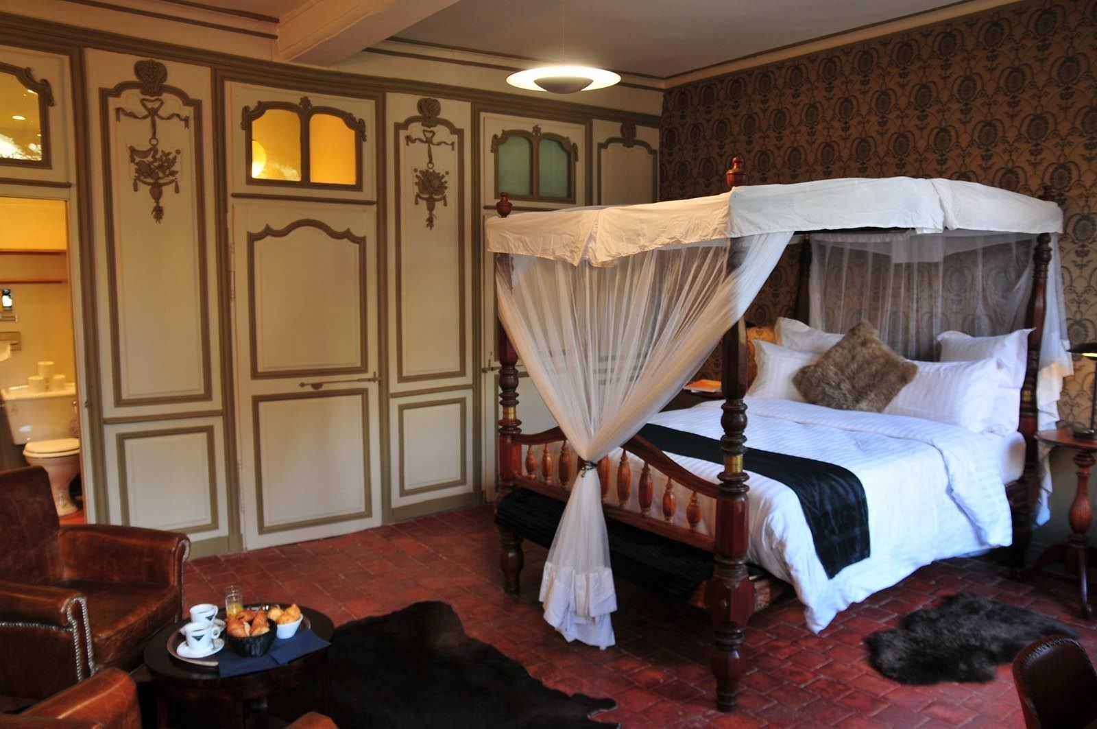 Een hemelbed met een wit beddegoed en kussens, een houten hoofdbord, en een rood vloerkleed op een rode baksteen vloer.