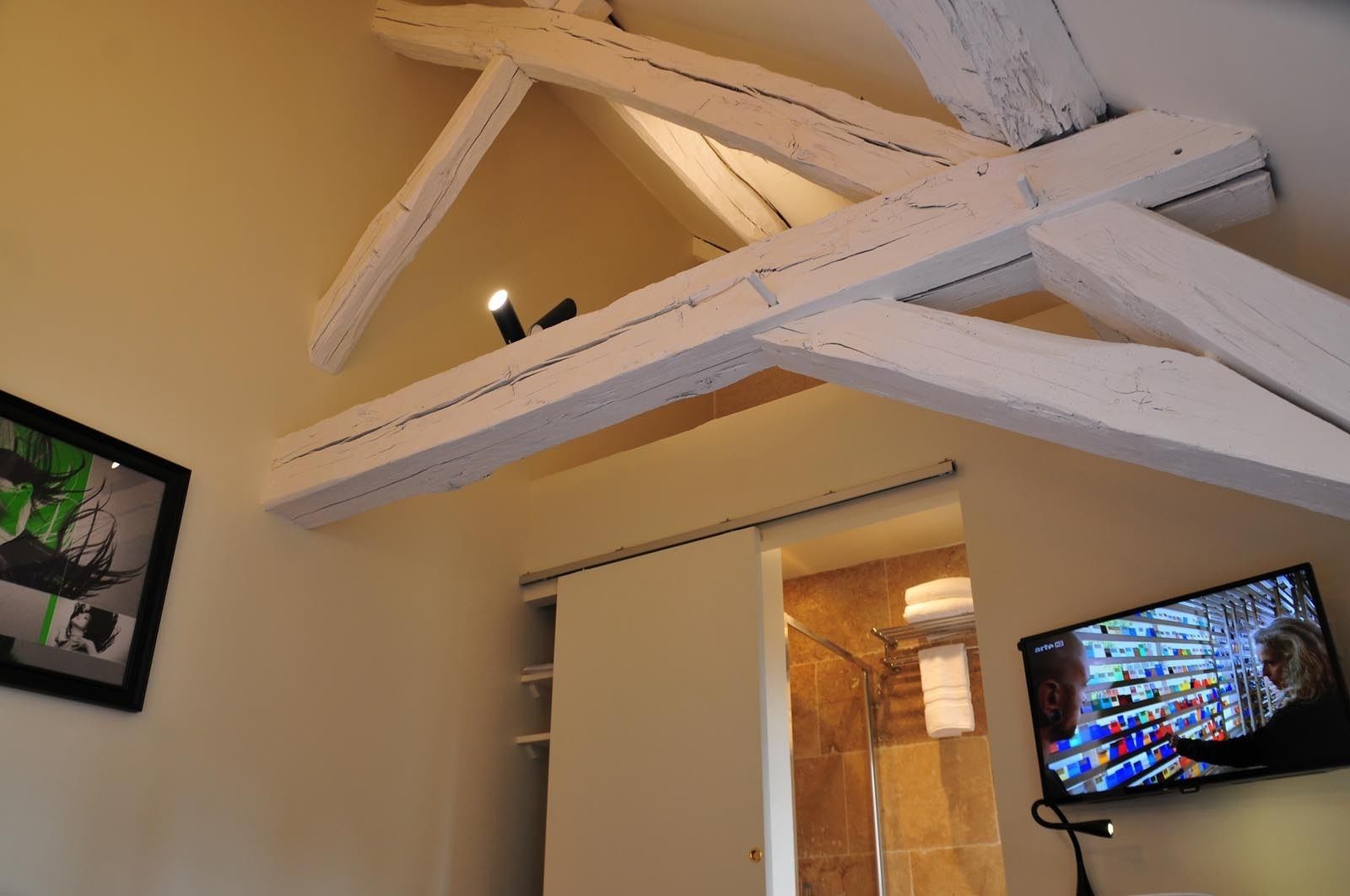 Quarto com vigas de madeira aparentes, área de chuveiro e televisão presa à parede.