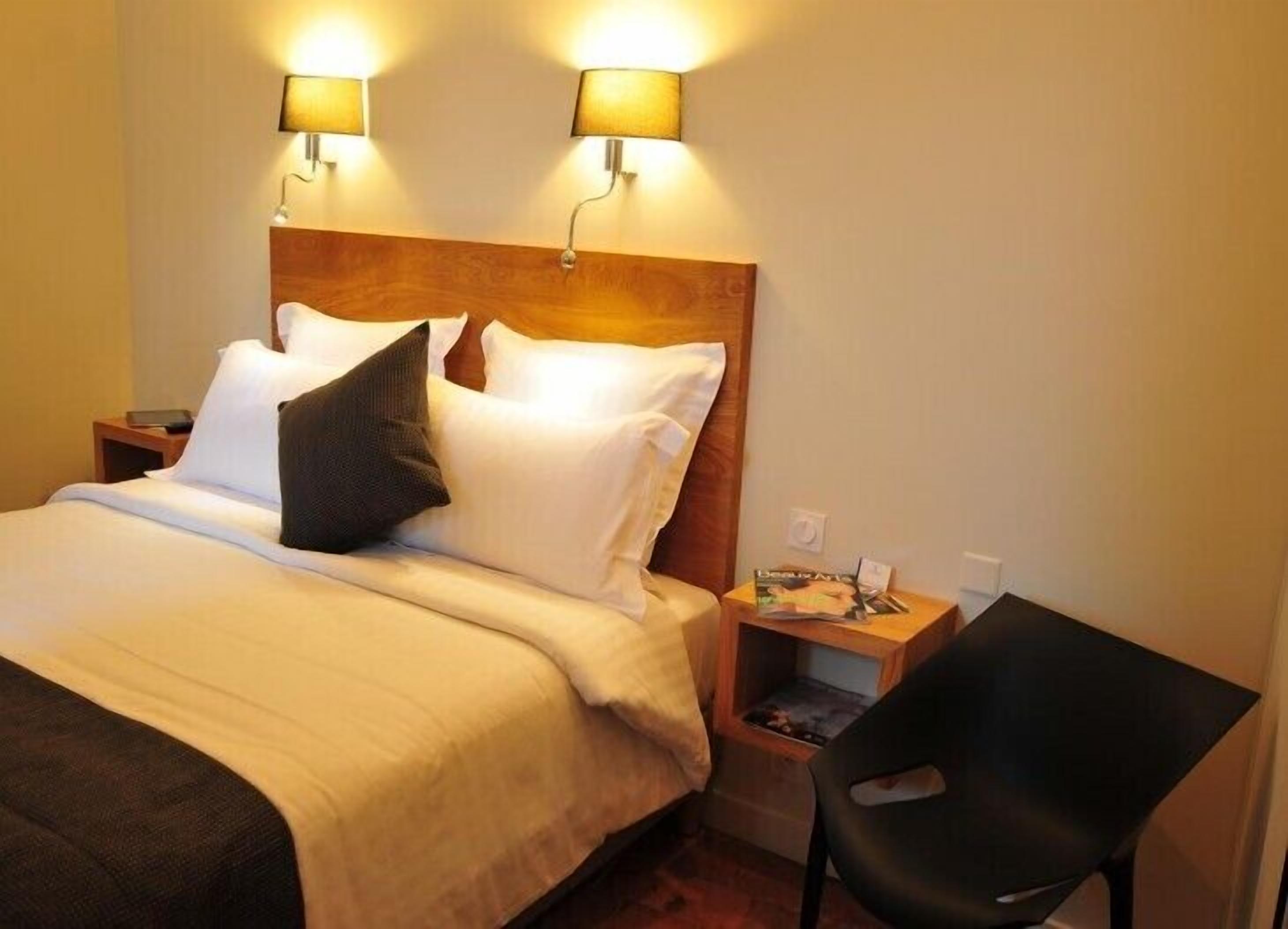 A hotel room with a wooden headboard, two bedside lamps, a bed with white linens, a black chair, and a small table with a magazine.