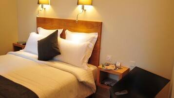 A hotel room with a wooden headboard, two bedside lamps, a bed with white linens, a black chair, and a small table with a magazine.