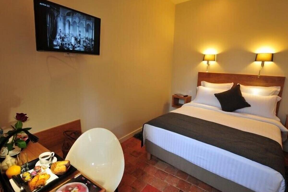 A hotel room with a bed, bedside lamps, a television on the wall, a small table with breakfast items, and a chair.