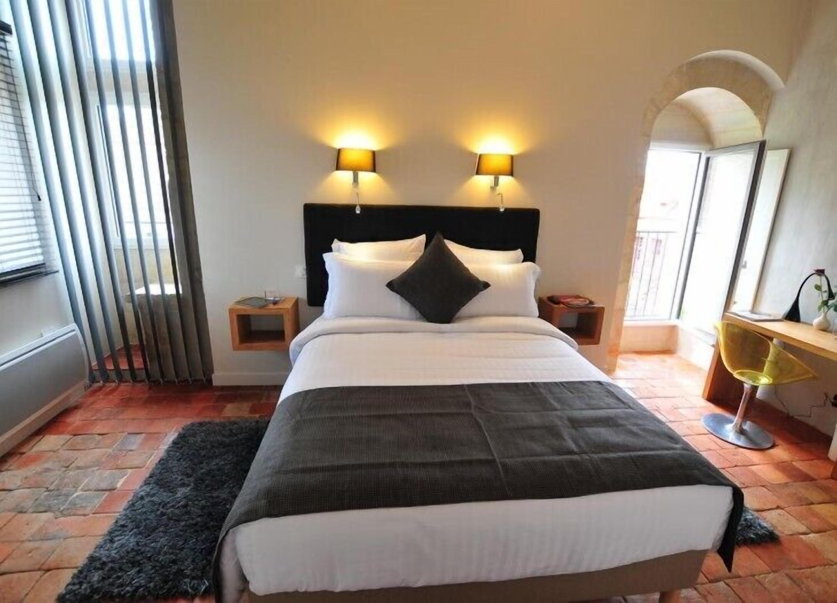 A neatly made bed with a black and white duvet, two bedside lamps, a small wooden nightstand, a black rug, and a yellow chair.