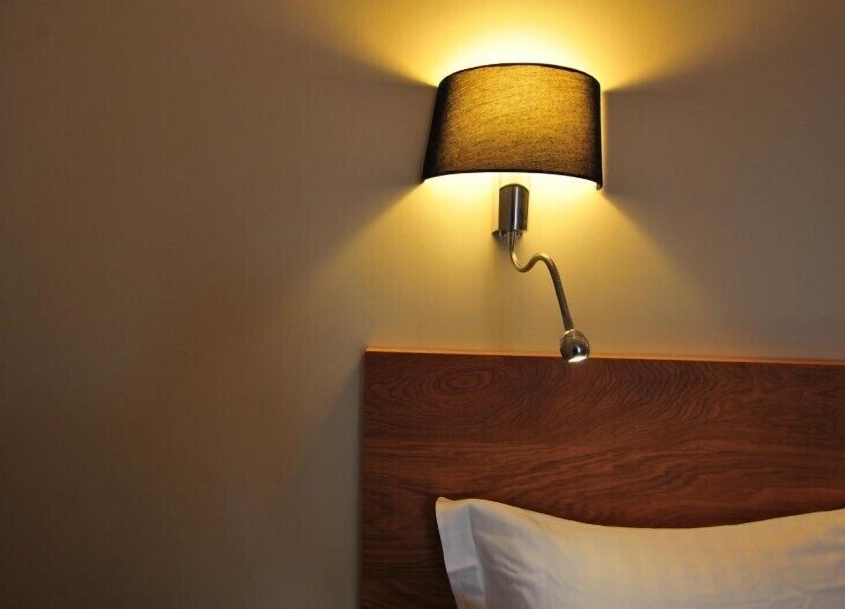 A wall-mounted lamp with a cylindrical shade illuminating a wooden headboard and a white pillow.