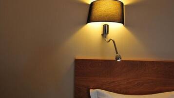 A wall-mounted lamp with a cylindrical shade illuminating a wooden headboard and a white pillow.