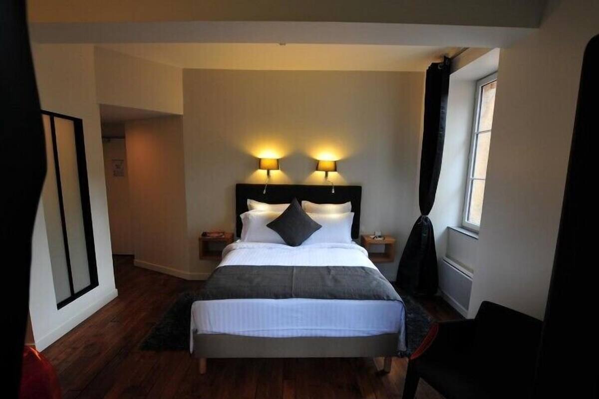 A neatly arranged hotel room with a large bed, two bedside lamps, a window with curtains, and a wooden floor.