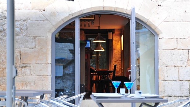 An outdoor seating area with metal chairs and tables, a stone wall, and a glass door leading to an interior space with a table, chairs, and a hanging light fixture. - Hôtel & Restaurant SY La Terrasse (Vezelay)